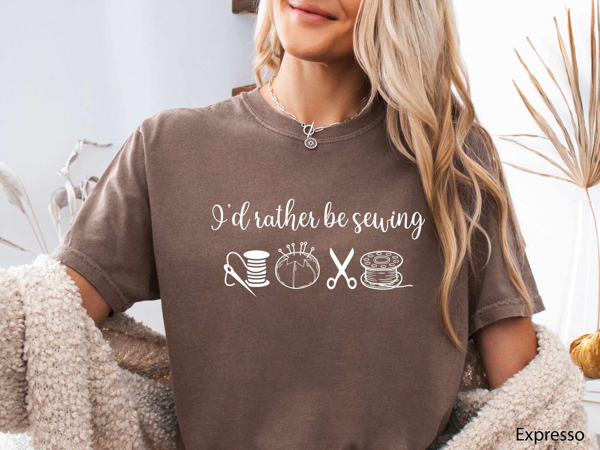 A t-shirt featuring the phrase “I’d Rather Be Sewing” with simple line-art icons of thread spools, a pincushion, scissors, and a needle and thread.
