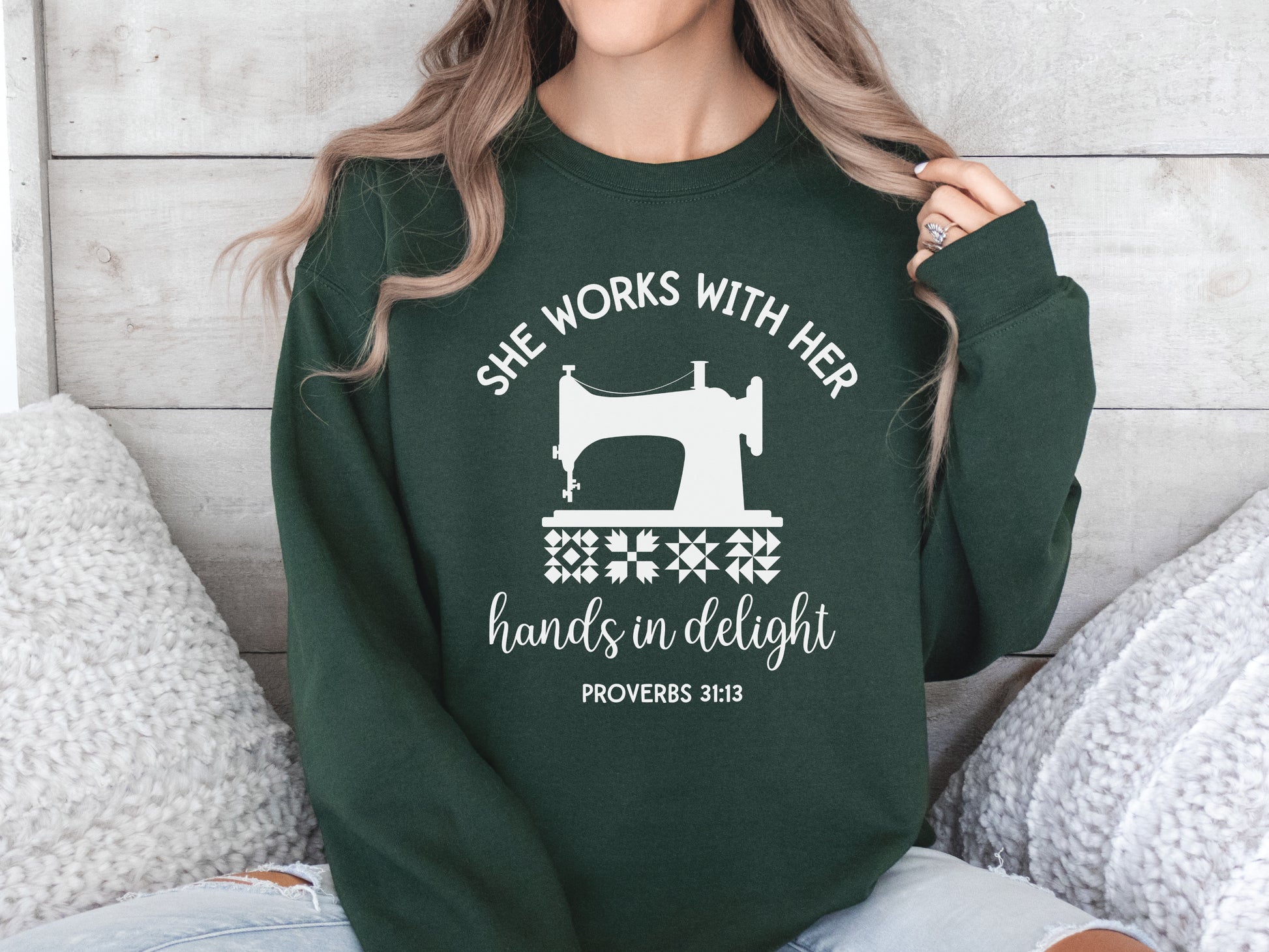 A crew style sweatshirt featuring a vintage-style sewing machine, quilt block icons, and the verse “She works with her hands in delight – Proverbs 31:13.”