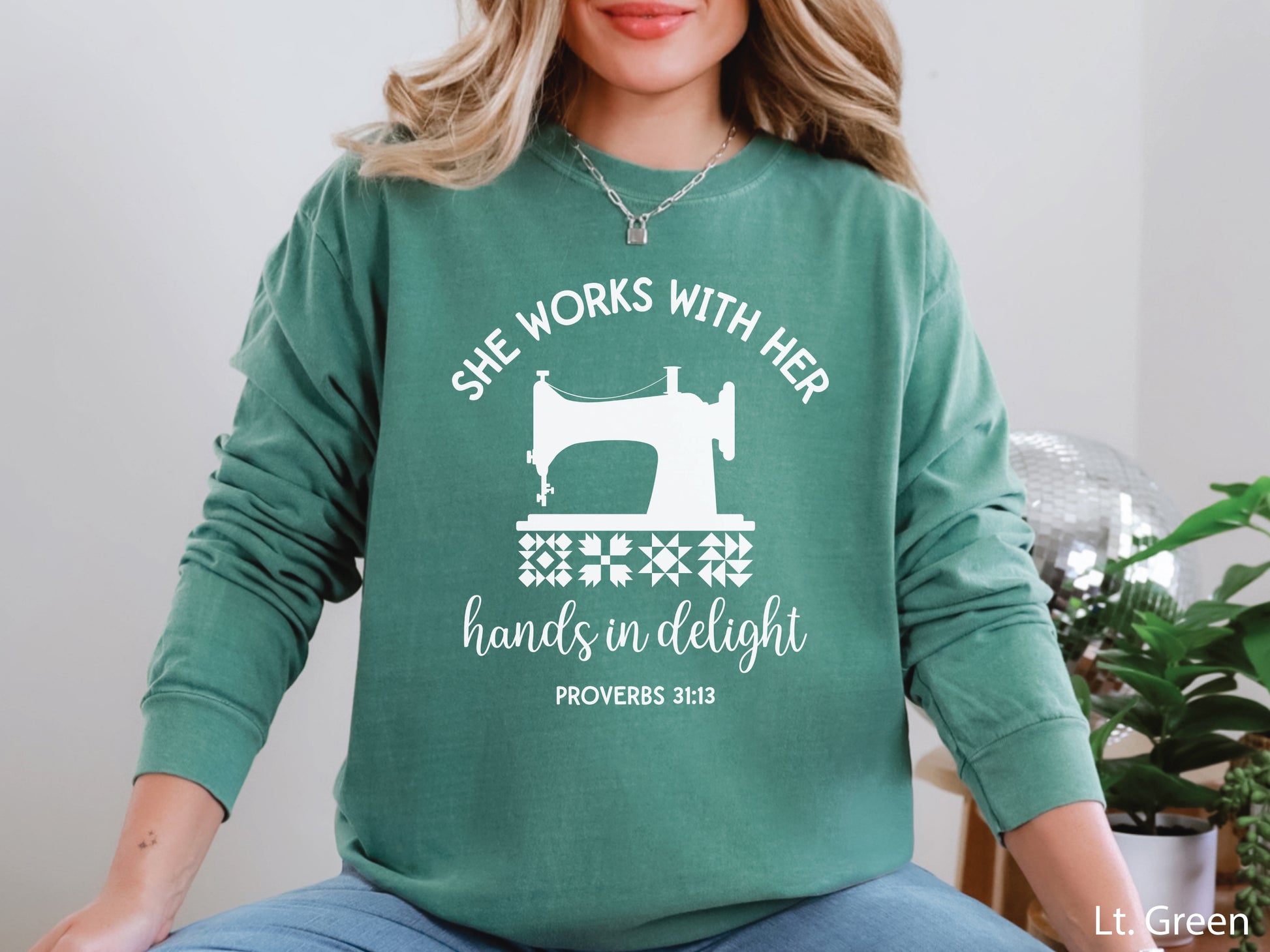 A long-sleeve  t-shirt featuring a vintage-style sewing machine, quilt block icons, and the verse “She works with her hands in delight – Proverbs 31:13.”