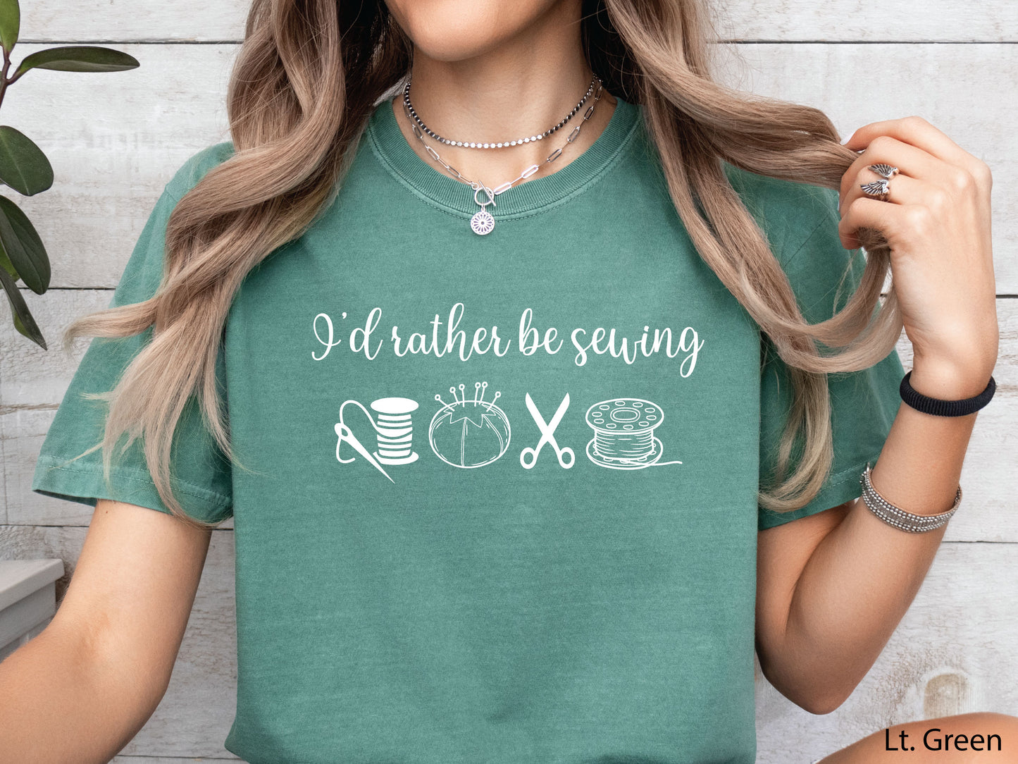 A t-shirt featuring the phrase “I’d Rather Be Sewing” with simple line-art icons of thread spools, a pincushion, scissors, and a needle and thread.
