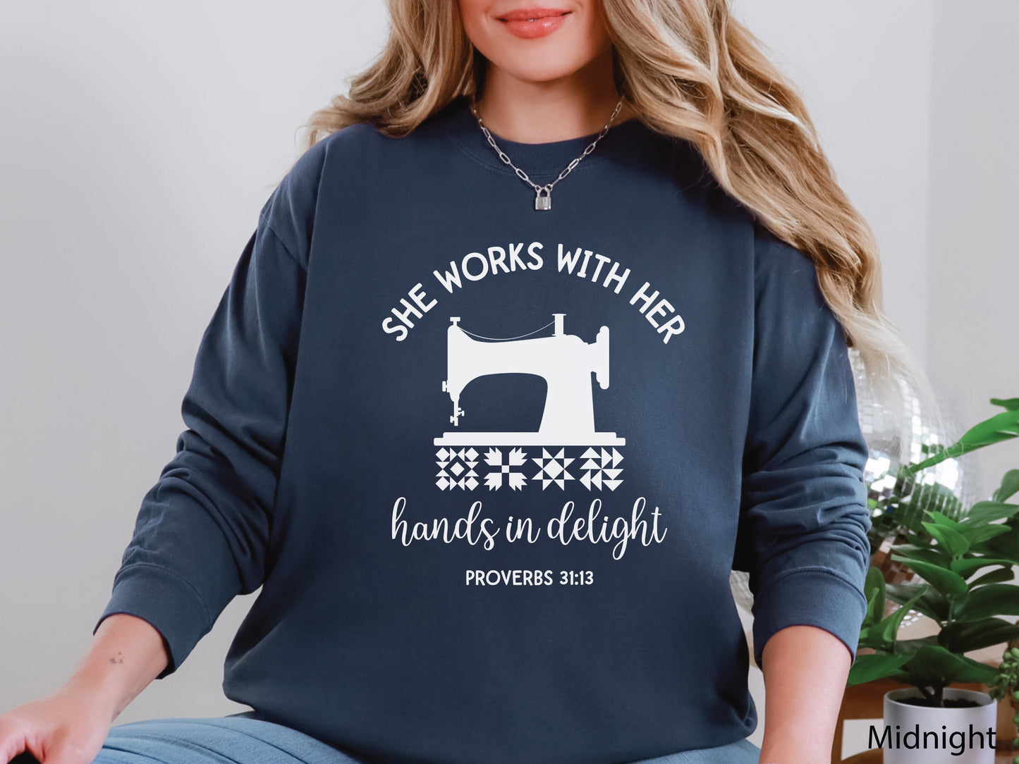 A long-sleeve  t-shirt featuring a vintage-style sewing machine, quilt block icons, and the verse “She works with her hands in delight – Proverbs 31:13.”