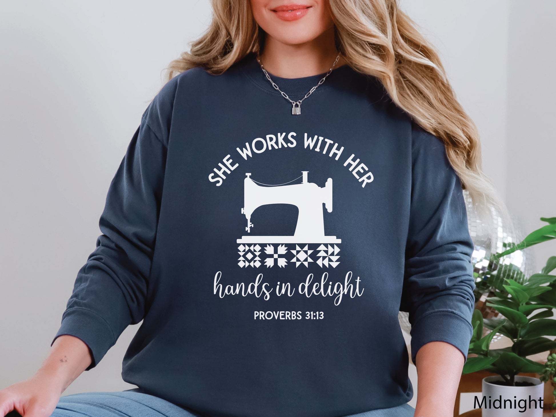 A long-sleeve  t-shirt featuring a vintage-style sewing machine, quilt block icons, and the verse “She works with her hands in delight – Proverbs 31:13.”