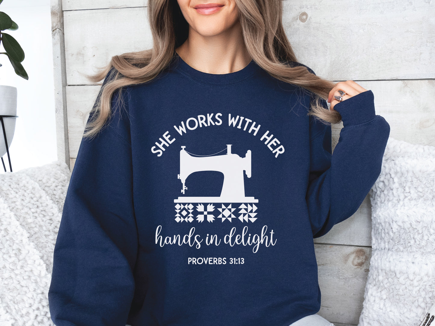 A crew style sweatshirt featuring a vintage-style sewing machine, quilt block icons, and the verse “She works with her hands in delight – Proverbs 31:13.”