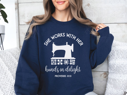 A crew style sweatshirt featuring a vintage-style sewing machine, quilt block icons, and the verse “She works with her hands in delight – Proverbs 31:13.”