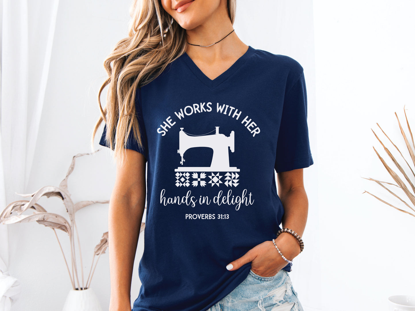 A quilt-themed v-neck  t-shirt featuring a vintage-style sewing machine, quilt block icons, and the verse “She works with her hands in delight – Proverbs 31:13.” 
