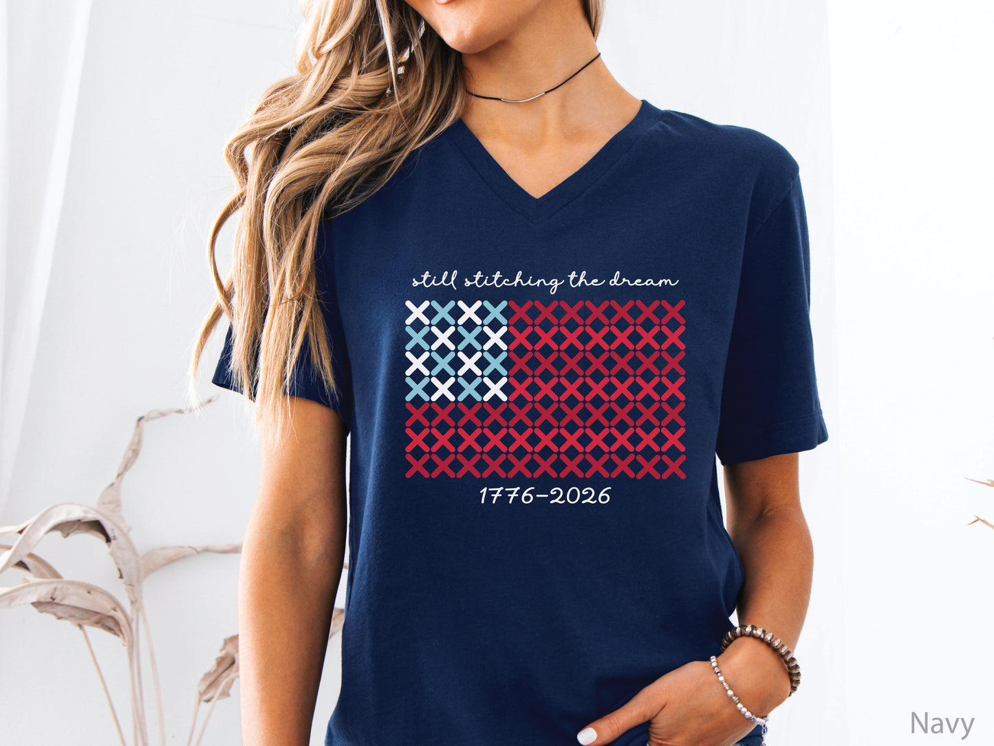 Patriotic v-neck t-shirt with cross-stitch American flag design and the phrase Still Stitching the Dream 1776–2026, ideal for quilters and needlework enthusiasts.