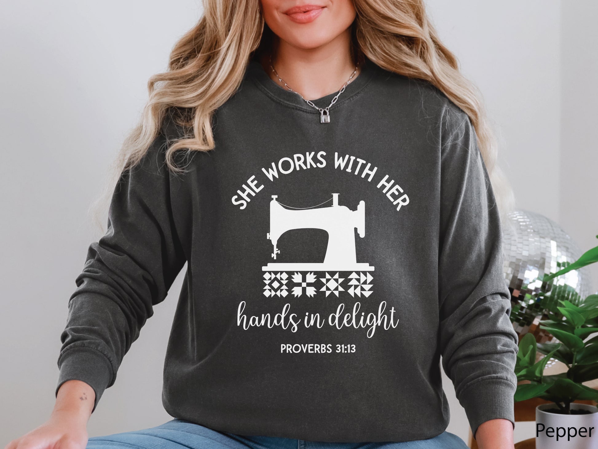 A long-sleeve  t-shirt featuring a vintage-style sewing machine, quilt block icons, and the verse “She works with her hands in delight – Proverbs 31:13.”
