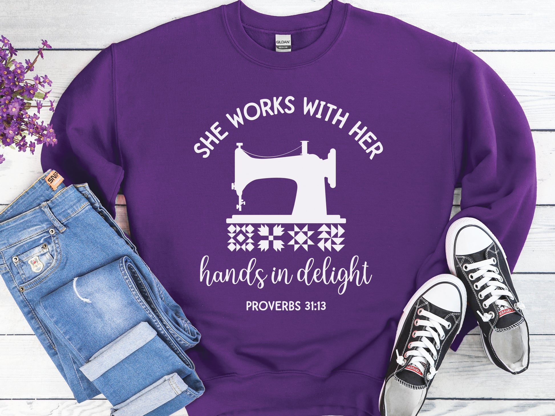 A crew style sweatshirt featuring a vintage-style sewing machine, quilt block icons, and the verse “She works with her hands in delight – Proverbs 31:13.”
