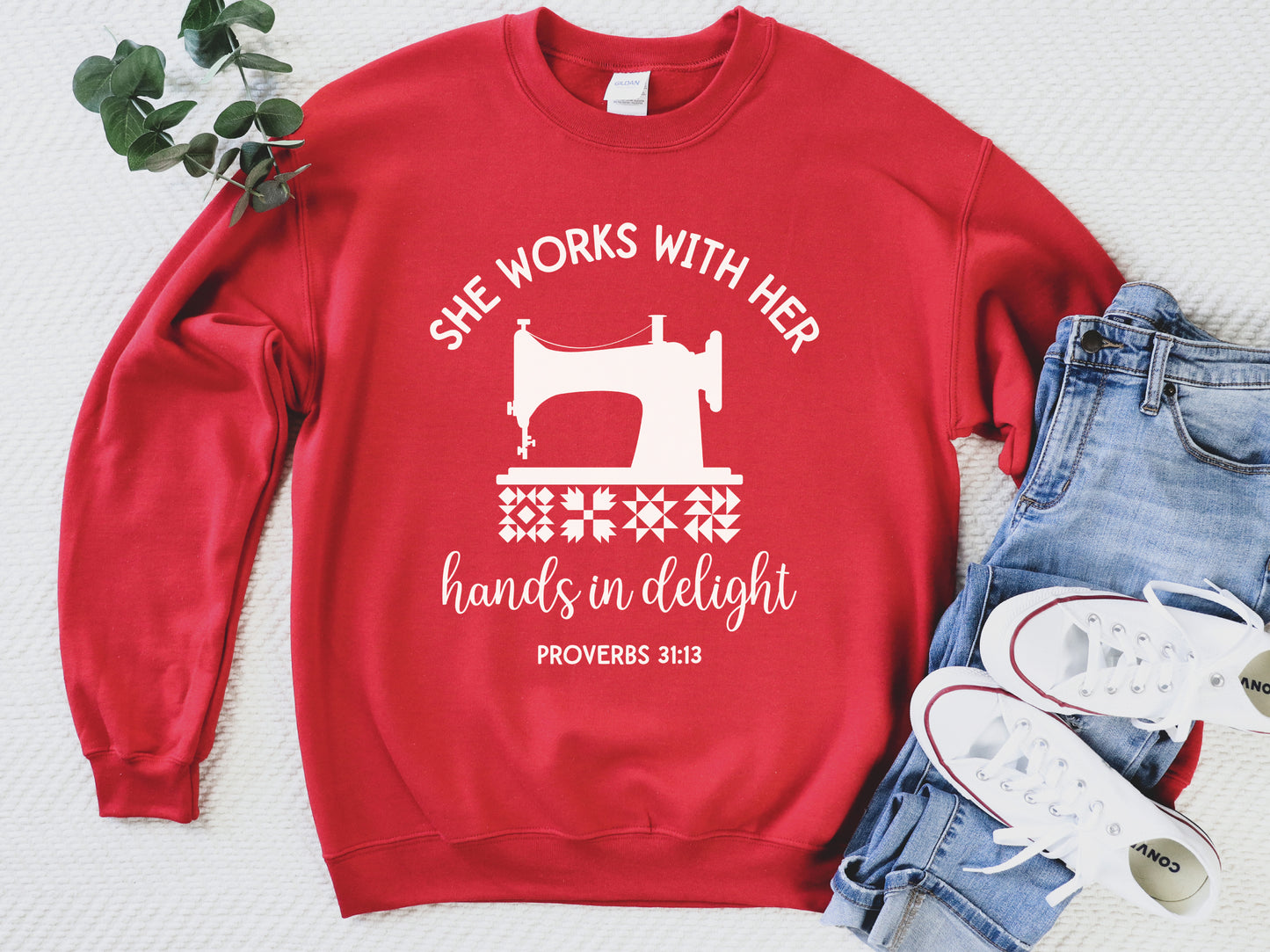 A crew style sweatshirt featuring a vintage-style sewing machine, quilt block icons, and the verse “She works with her hands in delight – Proverbs 31:13.”