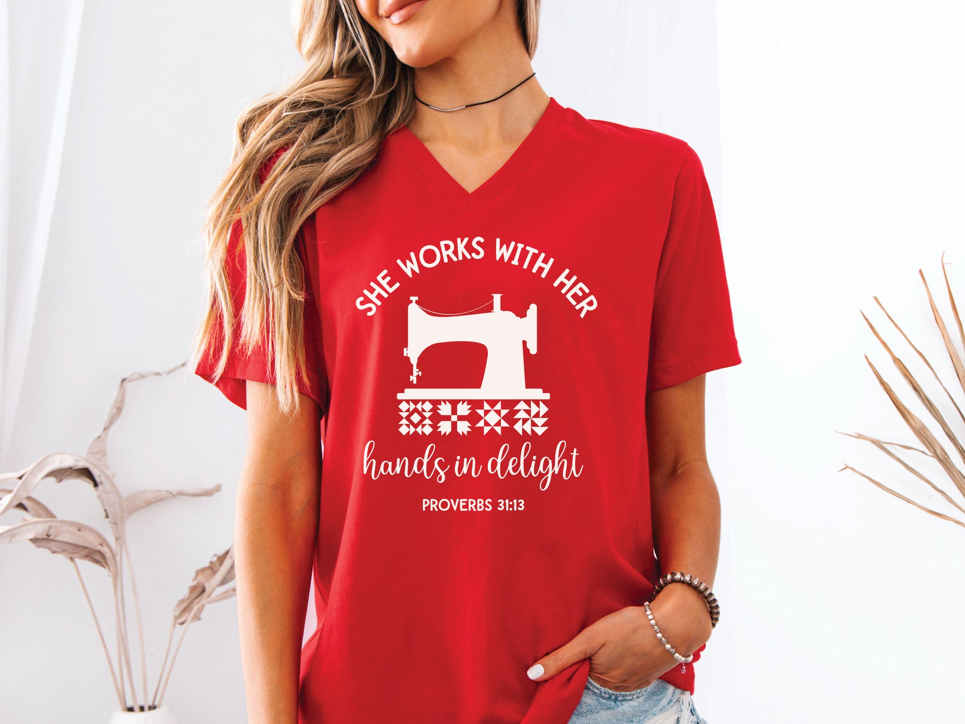 A quilt-themed v-neck  t-shirt featuring a vintage-style sewing machine, quilt block icons, and the verse “She works with her hands in delight – Proverbs 31:13.” 