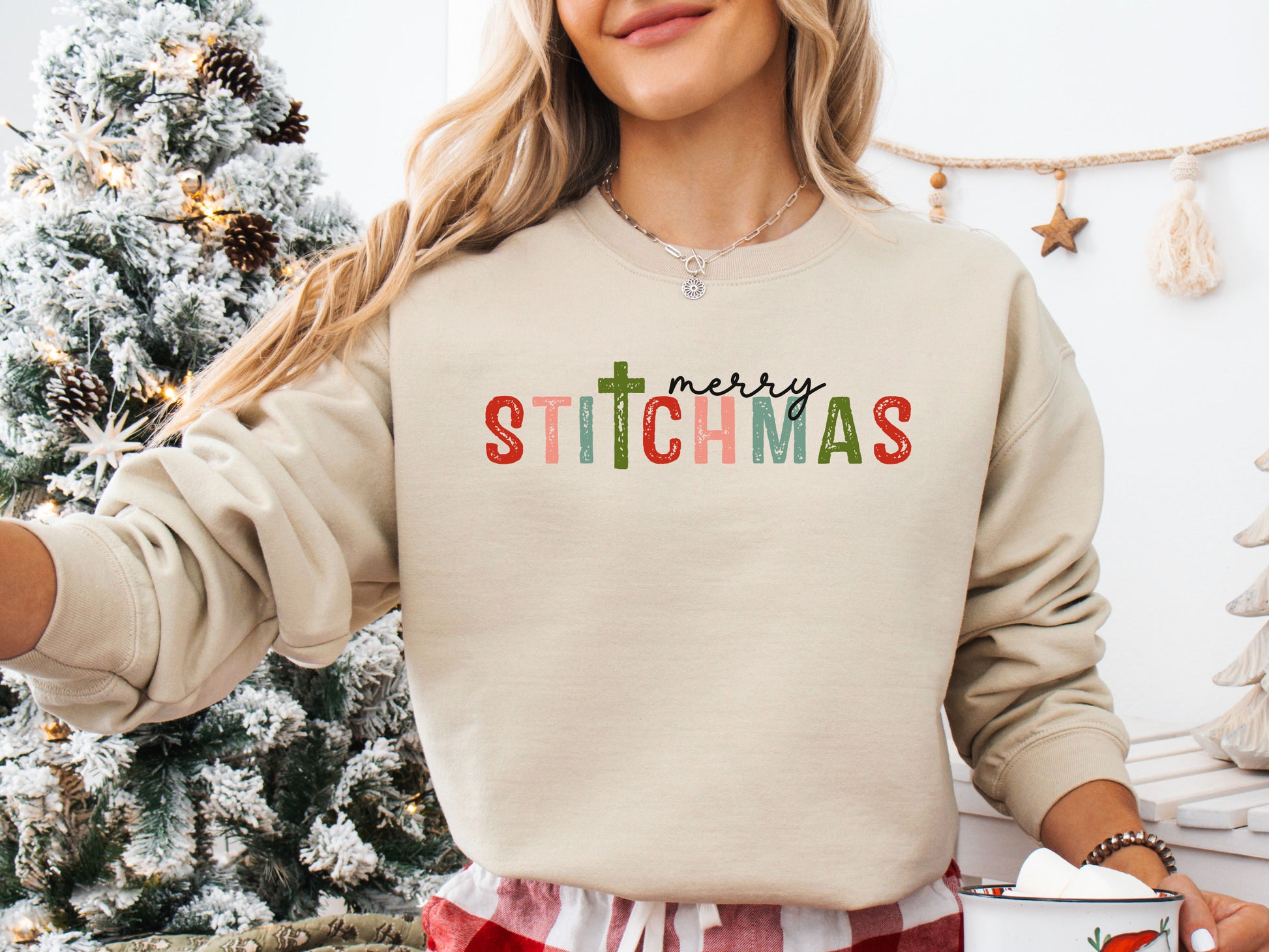 Beige (Sand) colored long sleeve shirt with the phrase “Merry Stitchmas” featuring a cross-shaped “T,” styled in a cozy Christmas setting with festive décor and sewing details.
