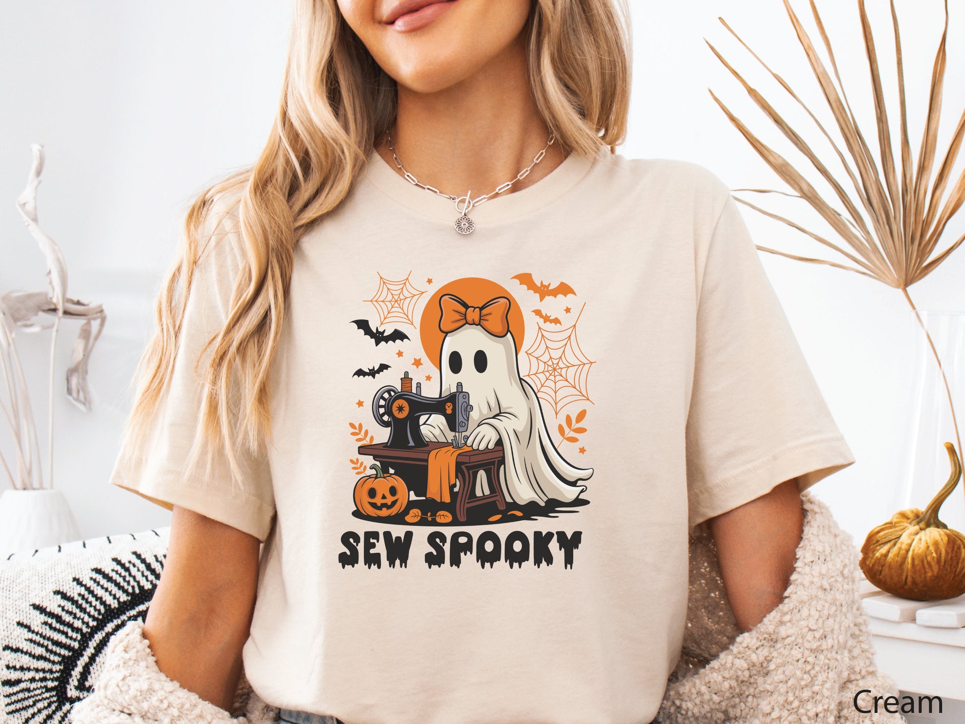 Cream color t-shirt with a Halloween sewing design featuring a ghost at a vintage sewing machine, surrounded by pumpkins, bats, spiderwebs, and stars, with the phrase “Sew Spooky” in bold black lettering.