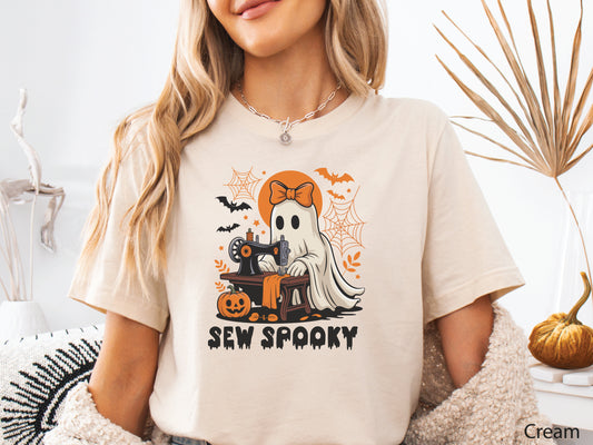 Cream color t-shirt with a Halloween sewing design featuring a ghost at a vintage sewing machine, surrounded by pumpkins, bats, spiderwebs, and stars, with the phrase “Sew Spooky” in bold black lettering.