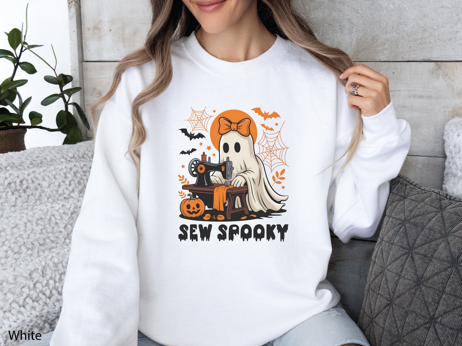 Cozy White sweatshirt with a Halloween sewing design featuring a ghost at a vintage sewing machine, surrounded by pumpkins, bats, spiderwebs, and stars, with the phrase “Sew Spooky” in bold black lettering.