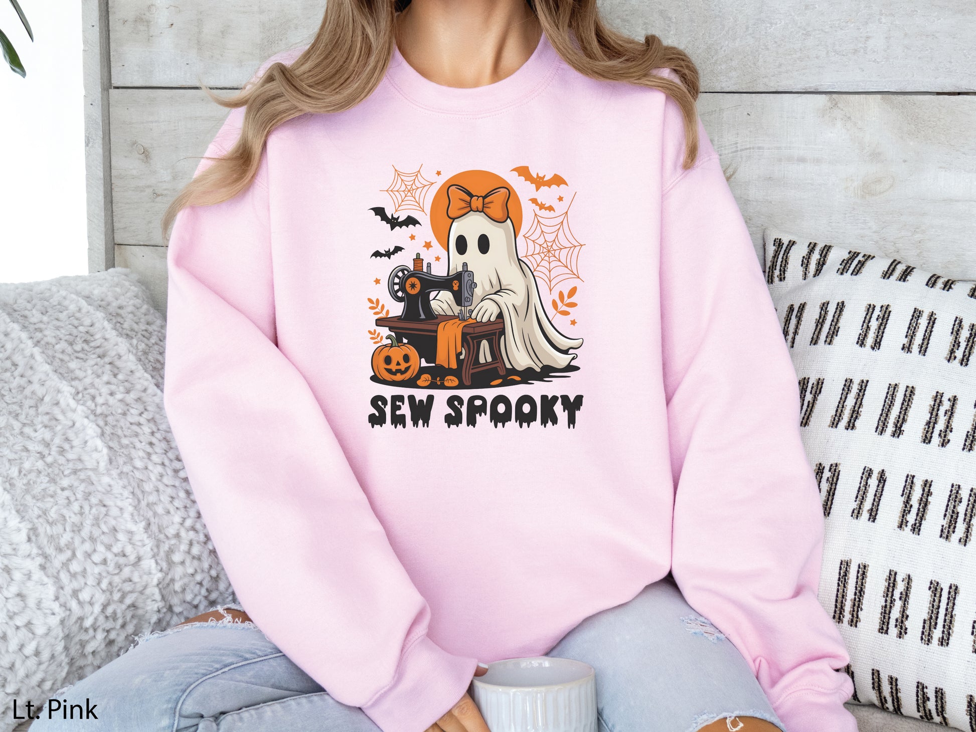 Cozy beige light pink color sweatshirt with a Halloween sewing design featuring a ghost at a vintage sewing machine, surrounded by pumpkins, bats, spiderwebs, and stars, with the phrase “Sew Spooky” in bold black lettering.