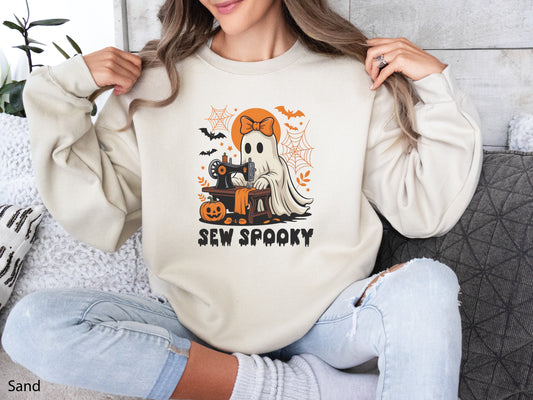 Cozy beige (Sand) color sweatshirt with a Halloween sewing design featuring a ghost at a vintage sewing machine, surrounded by pumpkins, bats, spiderwebs, and stars, with the phrase “Sew Spooky” in bold black lettering.