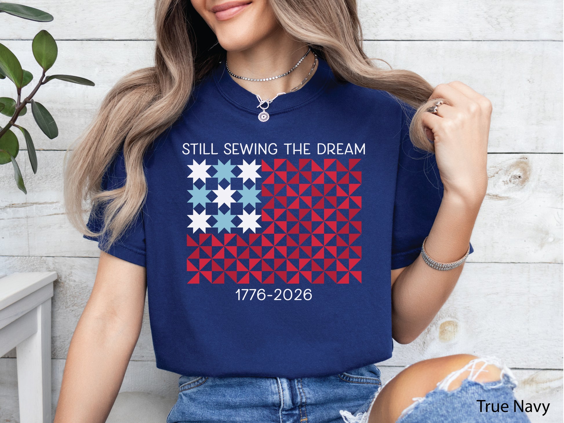 Patriotic quilting v-neck t-shirt with a quilt block American flag made of stars and pinwheel blocks and the phrase “Still Sewing the Dream 1776–2026” in red, white, and blue.