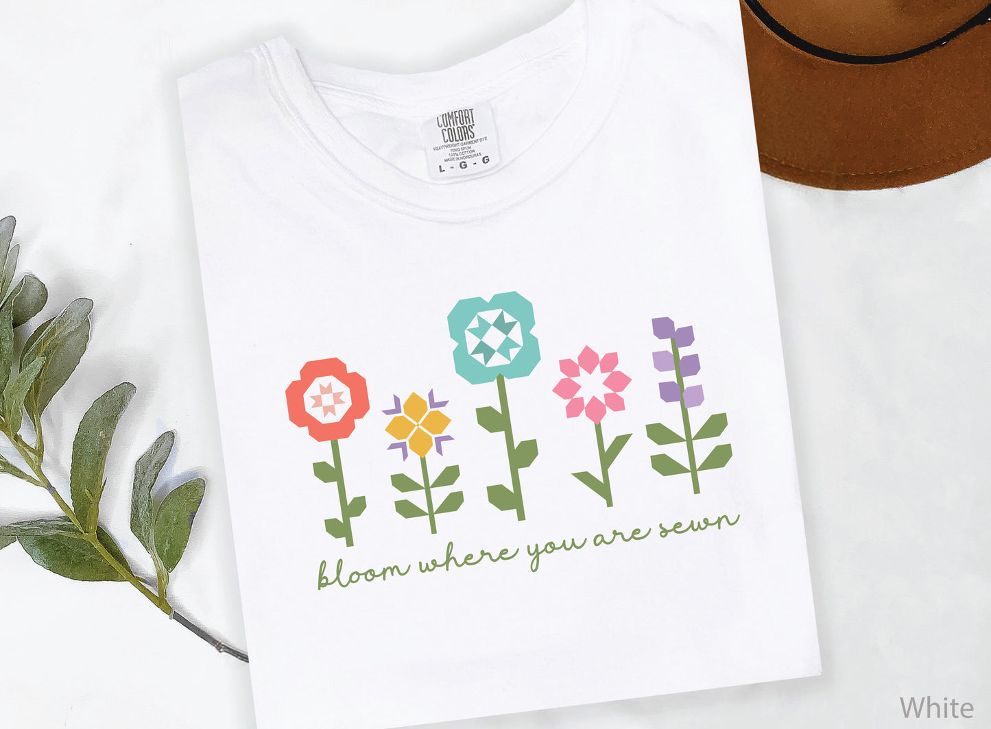 White soft t-shirt featuring colorful quilt block flowers with green stems and the phrase "Bloom Where You Are Sewn," perfect for quilters and sewing enthusiasts.