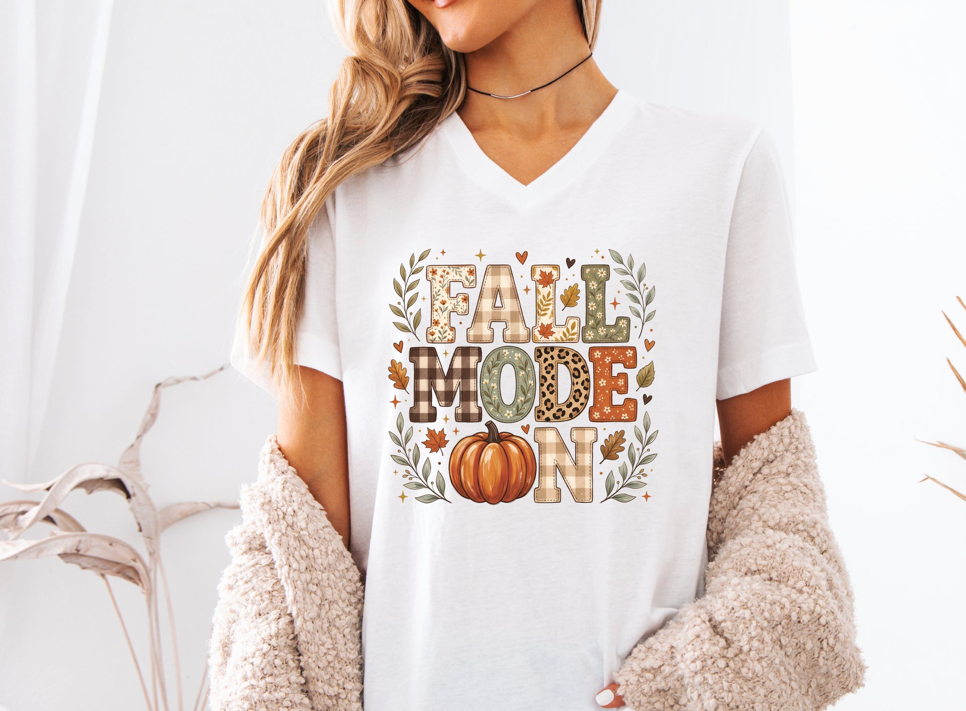 Fall Mode On v-neck t-shirt design featuring patchwork-style lettering with patchwork plaid, floral, leopard print, and fall leaf patterns, plus an orange pumpkin and autumn foliage.