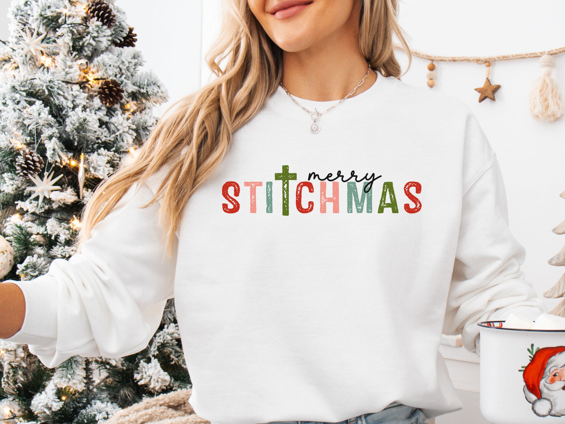 White colored long sleeve shirt with the phrase “Merry Stitchmas” featuring a cross-shaped “T,” styled in a cozy Christmas setting with festive décor and sewing details.