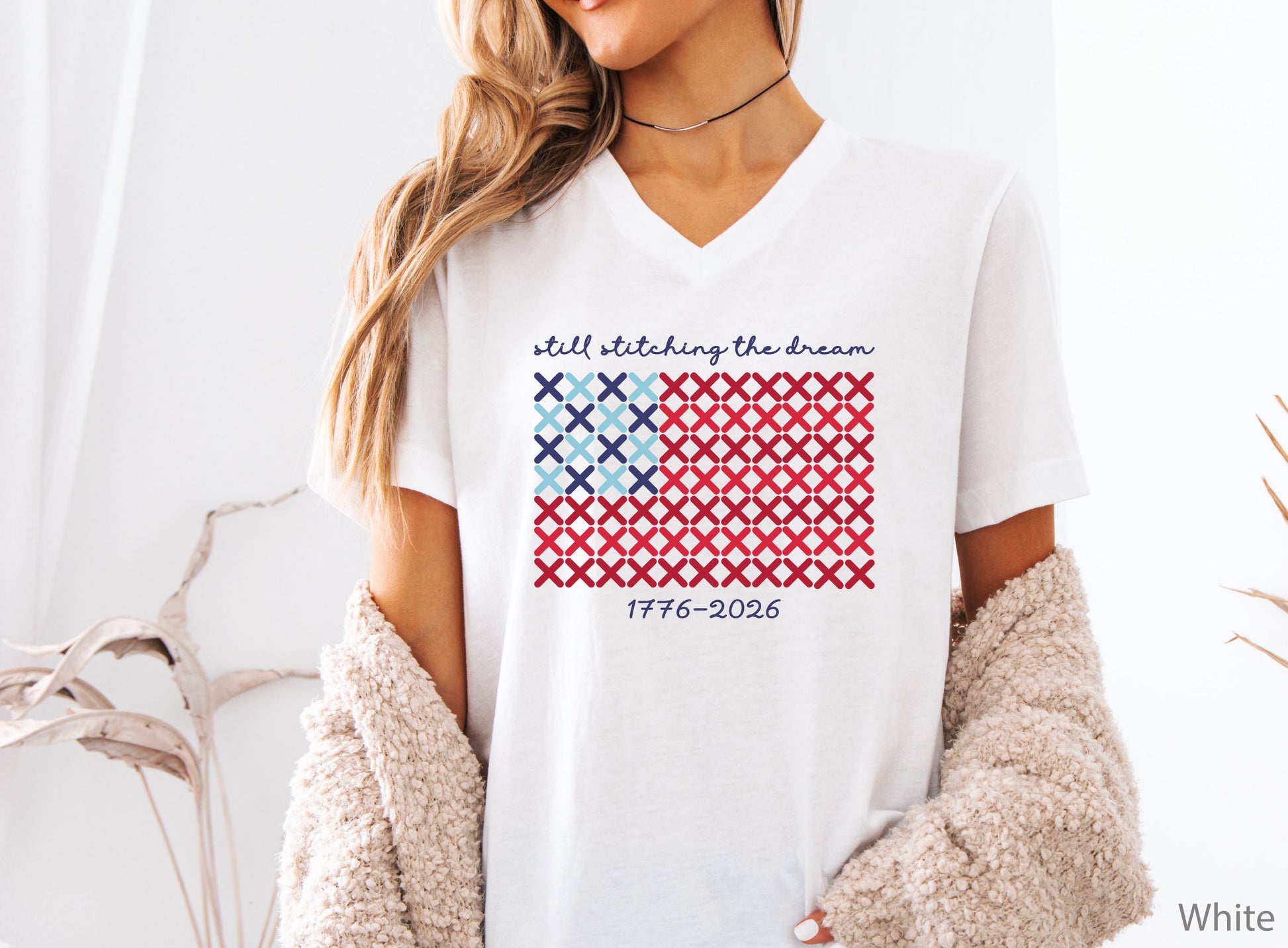 Patriotic v-neck t-shirt with cross-stitch American flag design and the phrase Still Stitching the Dream 1776–2026, ideal for quilters and needlework enthusiasts.
