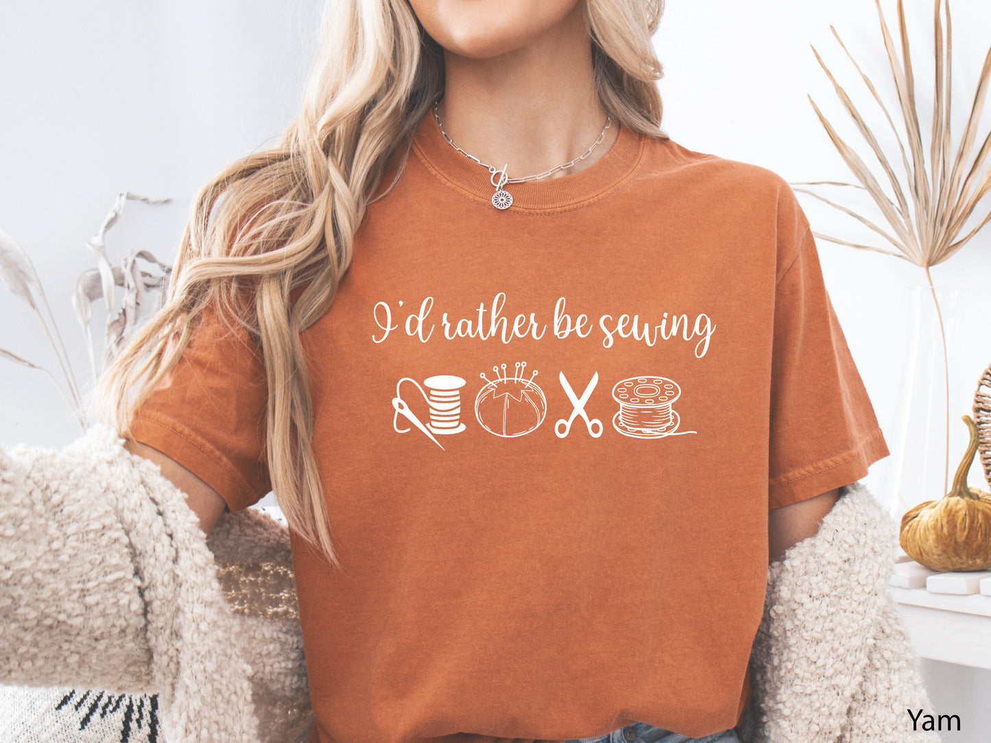 A t-shirt featuring the phrase “I’d Rather Be Sewing” with simple line-art icons of thread spools, a pincushion, scissors, and a needle and thread.